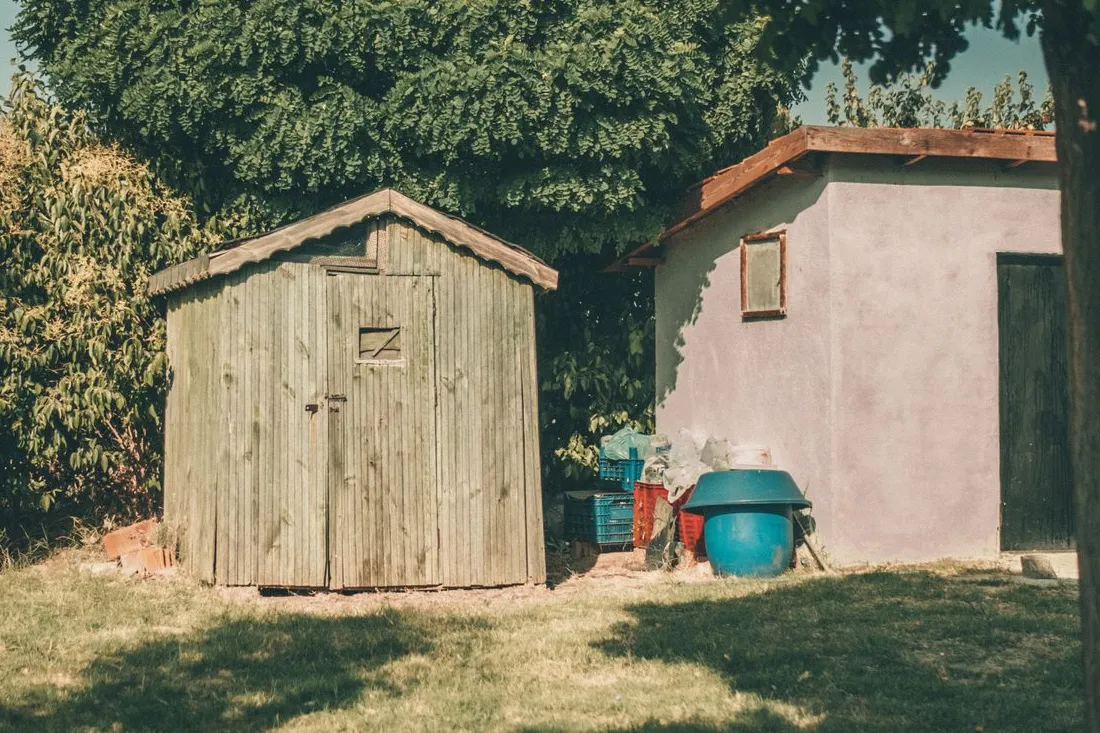 Petit abri de jardin en planches de bois grisées sur une pelouse, devant une haie, à côté d'une dépendance enduite